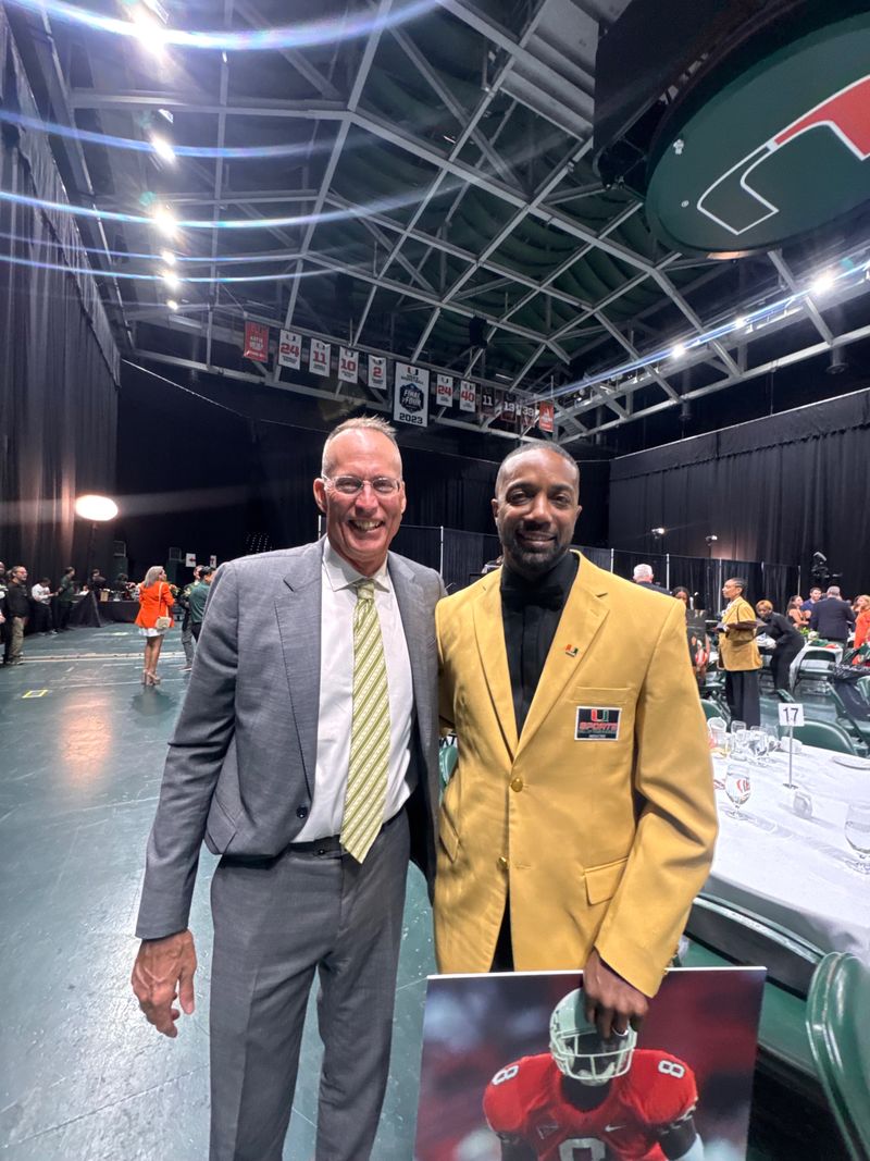 Mike Rumph, right, in his gold Hall of Fame jacket, poses with a guest inside the arena, holding a photo of a Miami player