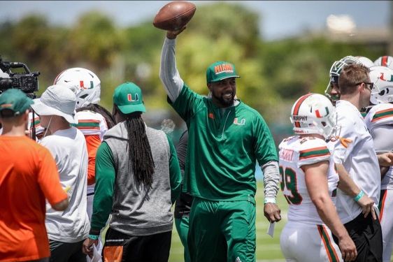 Mike Rumph, in green Miami staff gear and cap, holds a football overhead among Hurricanes players at practice