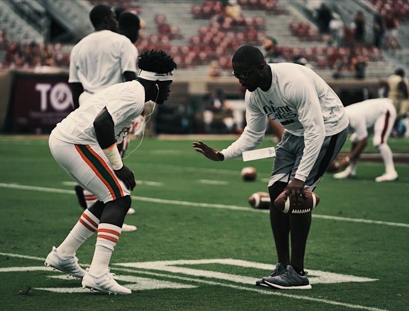 A coach in a Miami Canes shirt works with a Hurricanes defensive back during pregame warmups