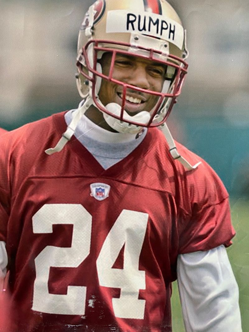 Mike Rumph smiling in a 49ers practice jersey, number 24, with RUMPH taped across the front of his helmet