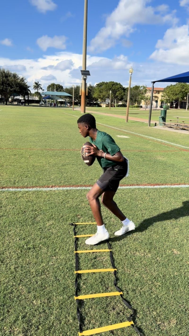 A young quarterback in a green shirt sets up with the football beside an agility ladder on a practice field