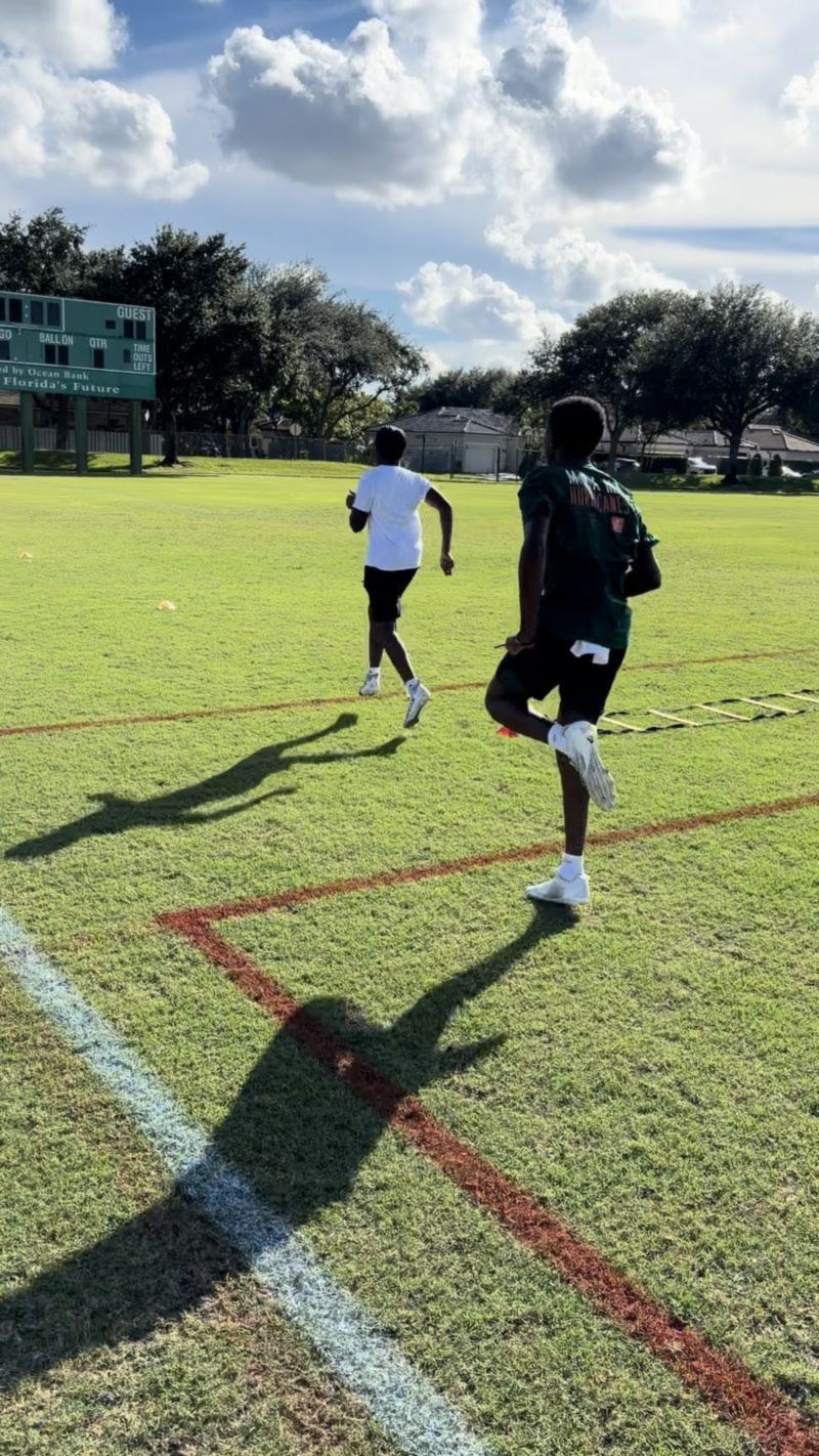 Two young athletes sprint across a sunny practice field past an agility ladder